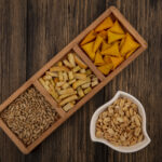 top view of cone shape bugles chips on a wooden divided plate with shelled sunflower seeds with pine nuts on a bowl on a wooden background